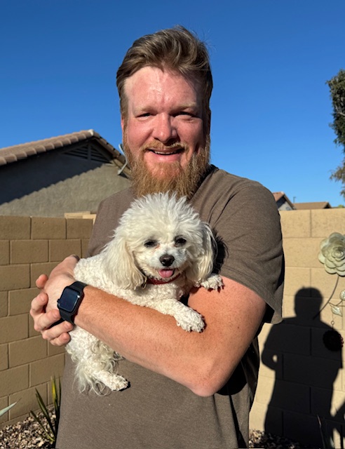 Adam Winston holding a small white dog outdoors on a sunny day, illustrating a calm moment of connection during trust-building visits with Meka the Bichon Frise.